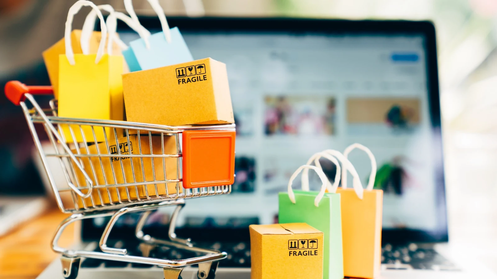 Carrinho de compras em miniatura cheio de caixas e sacolas coloridas sobre o teclado de um notebook com uma loja online aberta na tela