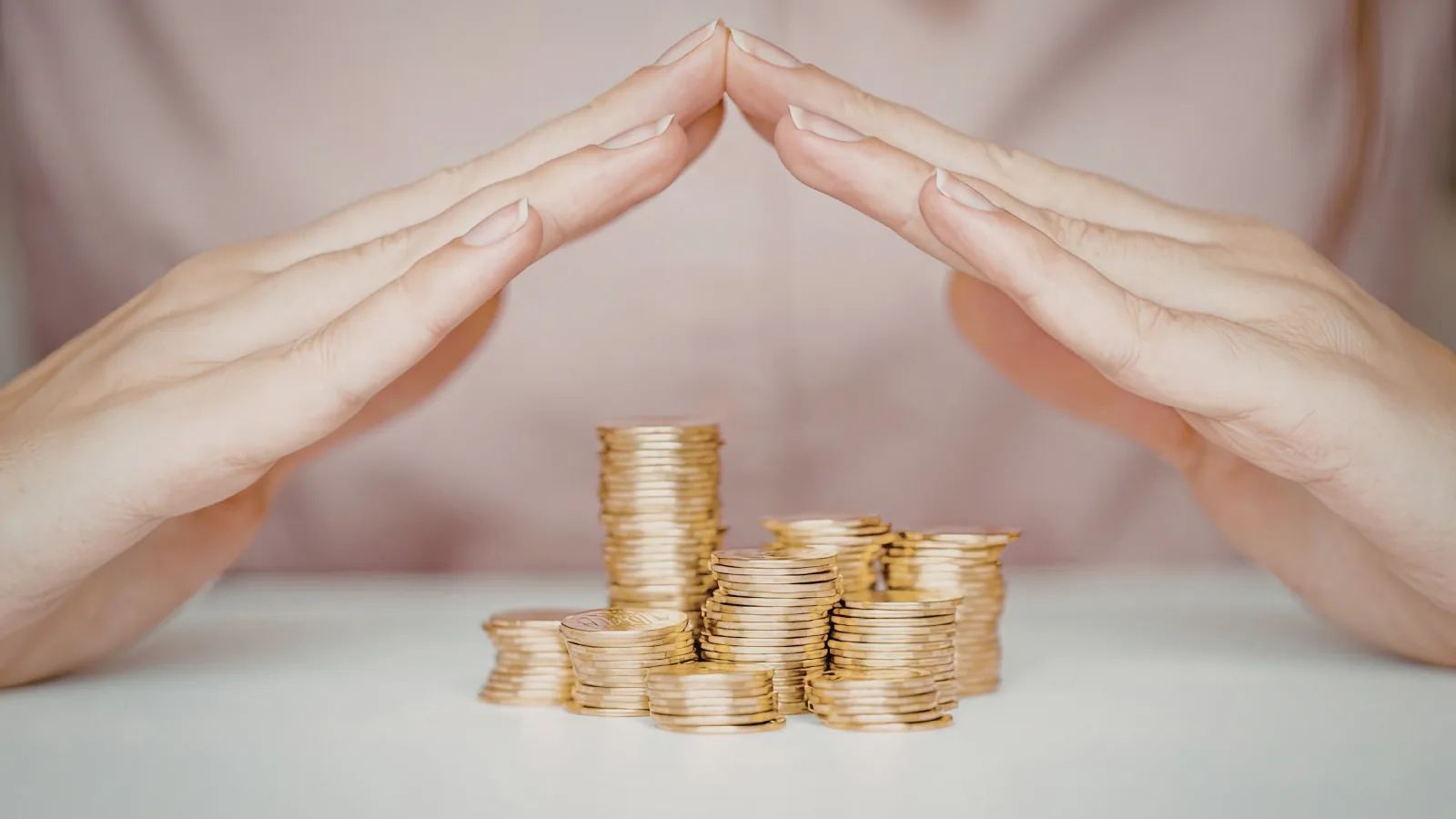 Mãos formando um telhado de proteção sobre pilhas de moedas douradas apoiadas numa superfície branca