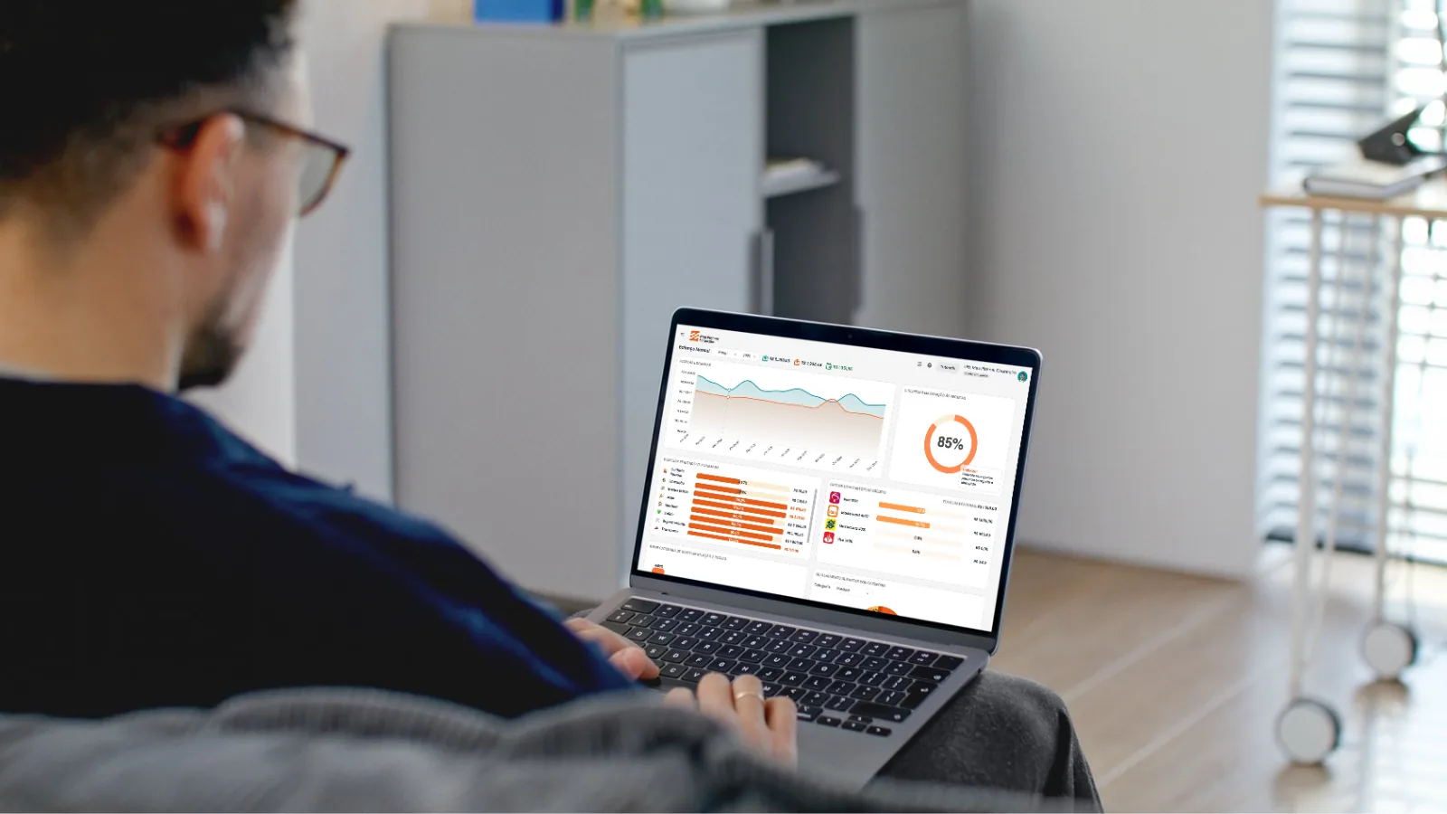 Homem de óculos, sentado no sofá com um notebook no colo, acompanha o balanço mensal das receitas e despesas no painel do Meu Planner Financeiro