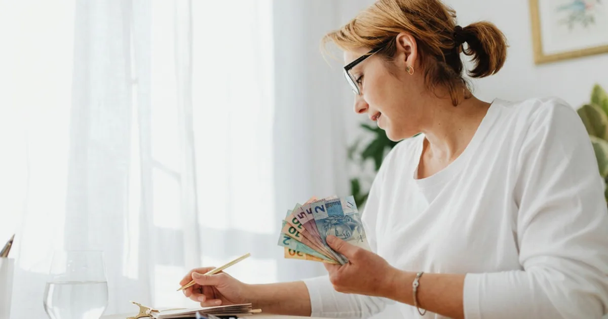 Mulher de óculos organizando as finanças pessoais, segurando cédulas de real com um caderno sobre a mesa